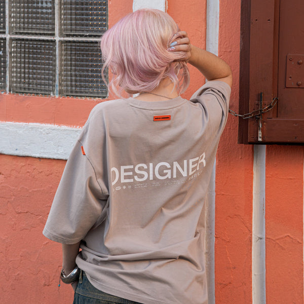 Person with pink hair wearing a beige t-shirt with 'DESIGNER' text against a red brick wall.